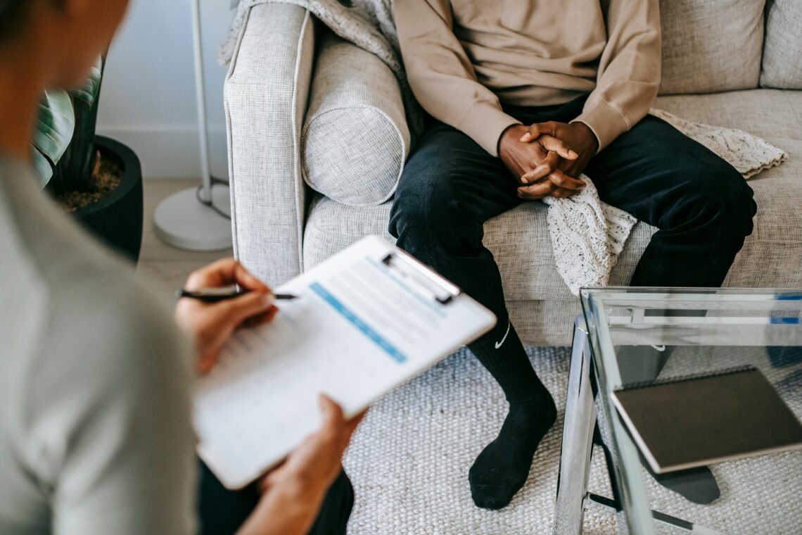 A doctor taking notes on a clipboard while consulting with a male patient sitting on a couch.