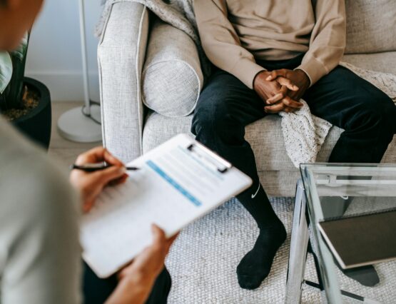 A doctor taking notes on a clipboard while consulting with a male patient sitting on a couch.